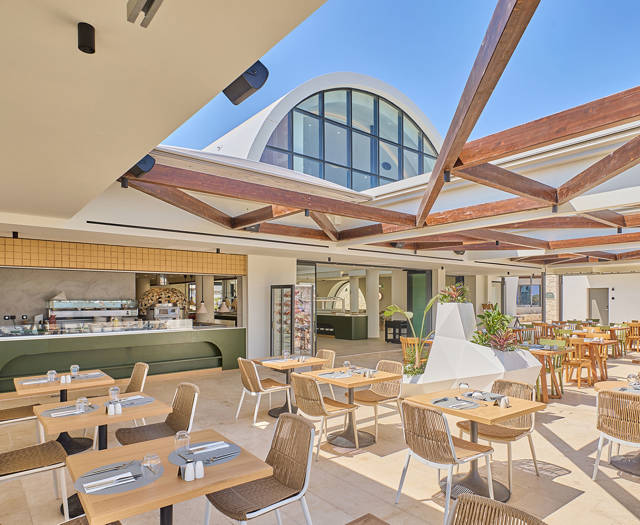 Bright and airy restaurant terrace with wooden beams, modern decor, and an open kitchen under a blue sky.