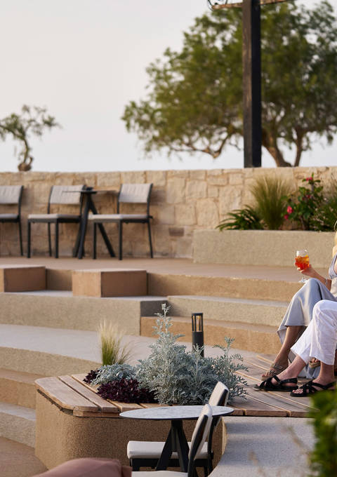Couple enjoying drinks while seated on an outdoor lounge overlooking terraced steps and greenery near the sea.