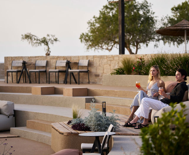Couple enjoying drinks while seated on an outdoor lounge overlooking terraced steps and greenery near the sea.