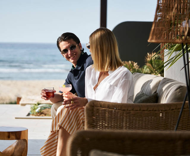 Couple seated on a wicker sofa by the beach, enjoying cocktails and smiling at each other.