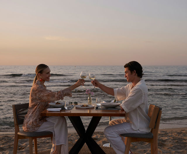 Couple sitting at a table on the beach during sunset, clinking wine glasses over a meal with the sea in the background.
