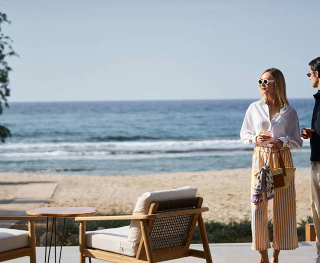Stylish couple enjoying drinks by the beach, standing near outdoor lounge chairs with a view of the ocean.