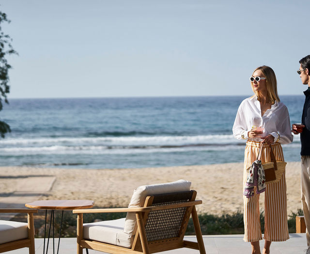 Stylish couple enjoying drinks by the beach, standing near outdoor lounge chairs with a view of the ocean.
