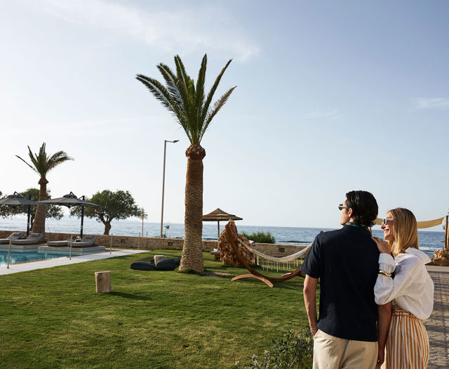 Couple enjoying sea view from the lawn at Kosta Alimia, with palm trees and hammocks nearby.