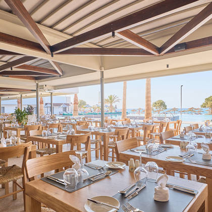 Seaside restaurant with wooden tables set for dining, offering views of palm trees and sunbeds by the water.
