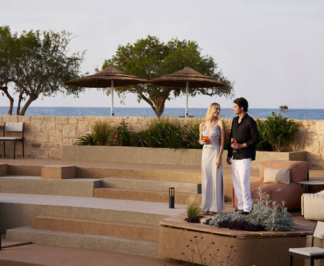 Couple dressed elegantly, standing and enjoying drinks in an outdoor terraced lounge area overlooking the sea with trees and sun umbrellas.
