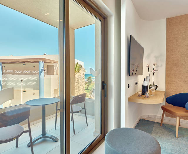 Side sea view balcony with table and chairs, seen from a bright room with desk and TV.