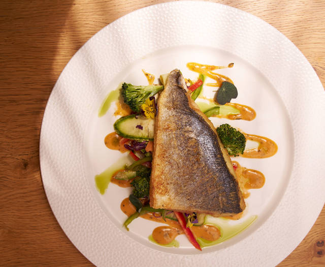 Seared fish fillet served over sautéed vegetables and drizzled with vibrant sauces on a white plate.