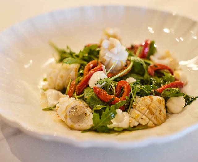 Grilled calamari salad with arugula, roasted red peppers, and citrus dressing in a scalloped white bowl.