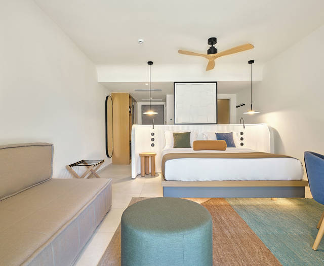 Bright junior suite with double bed, sofa, blue and green chairs, and colorful area rug.