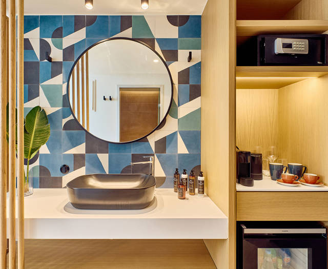 Modern bathroom vanity with round mirror, patterned wall tiles, and adjacent minibar with coffee station and safe.