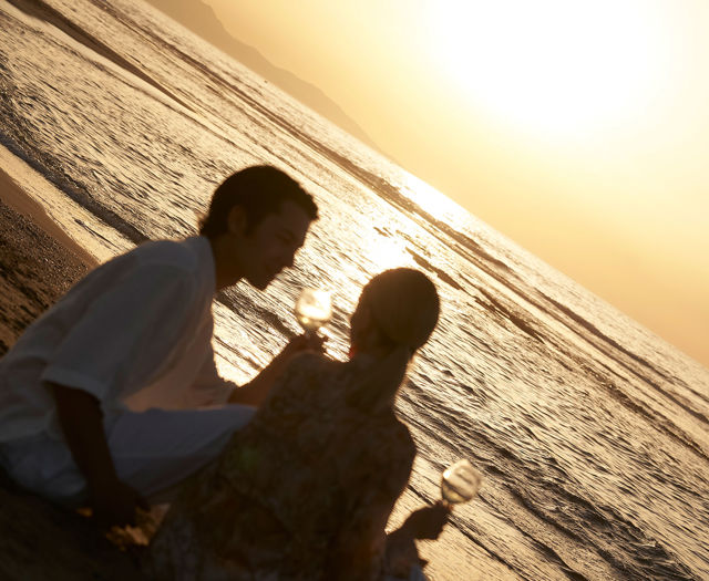 Couple sitting on beach at sunset holding wine glasses.