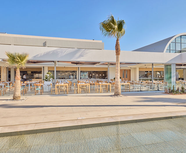 Bright daytime view of a stylish open-air restaurant with palm trees and a shallow reflecting pool in front.