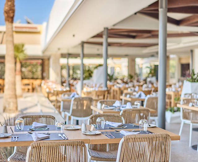 Elegant outdoor restaurant setup with wooden tables, woven chairs, and palm trees under a clear sky.