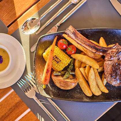 Grilled chop with roasted vegetables and potato wedges, served alongside a creamy soup garnished with a purple edible flower.