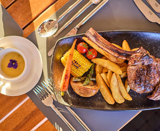 Grilled chop with roasted vegetables and potato wedges, served alongside a creamy soup garnished with a purple edible flower.
