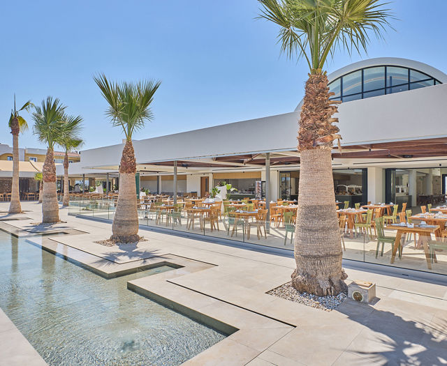 Modern restaurant with palm trees and tiered water features, set beneath a bright blue sky.