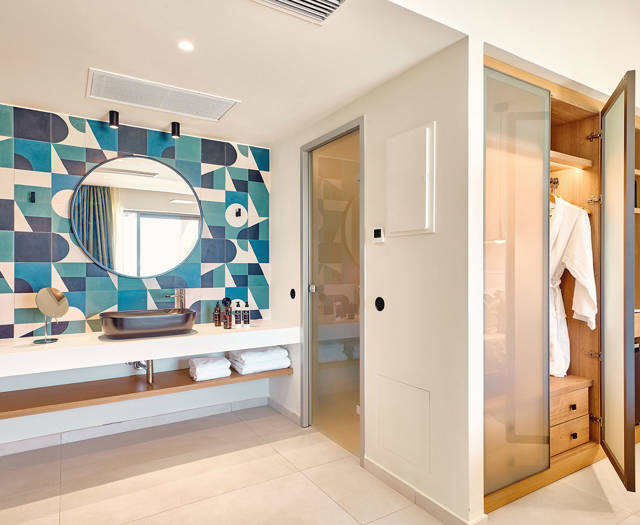 Double room's stylish bathroom with geometric tiled wall, round mirror, and open wardrobe with robes