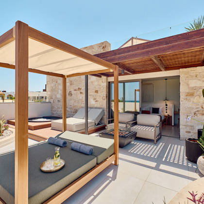 Sunlit terrace with canopy daybed, lounge chairs, and outdoor seating in front of a stone wall facade.