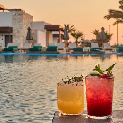 Two colorful cocktails garnished with herbs and berries placed on a table by the poolside at sunset.