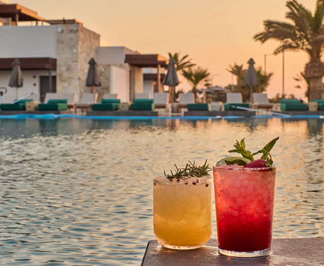 Two colorful cocktails garnished with herbs and berries placed on a table by the poolside at sunset.
