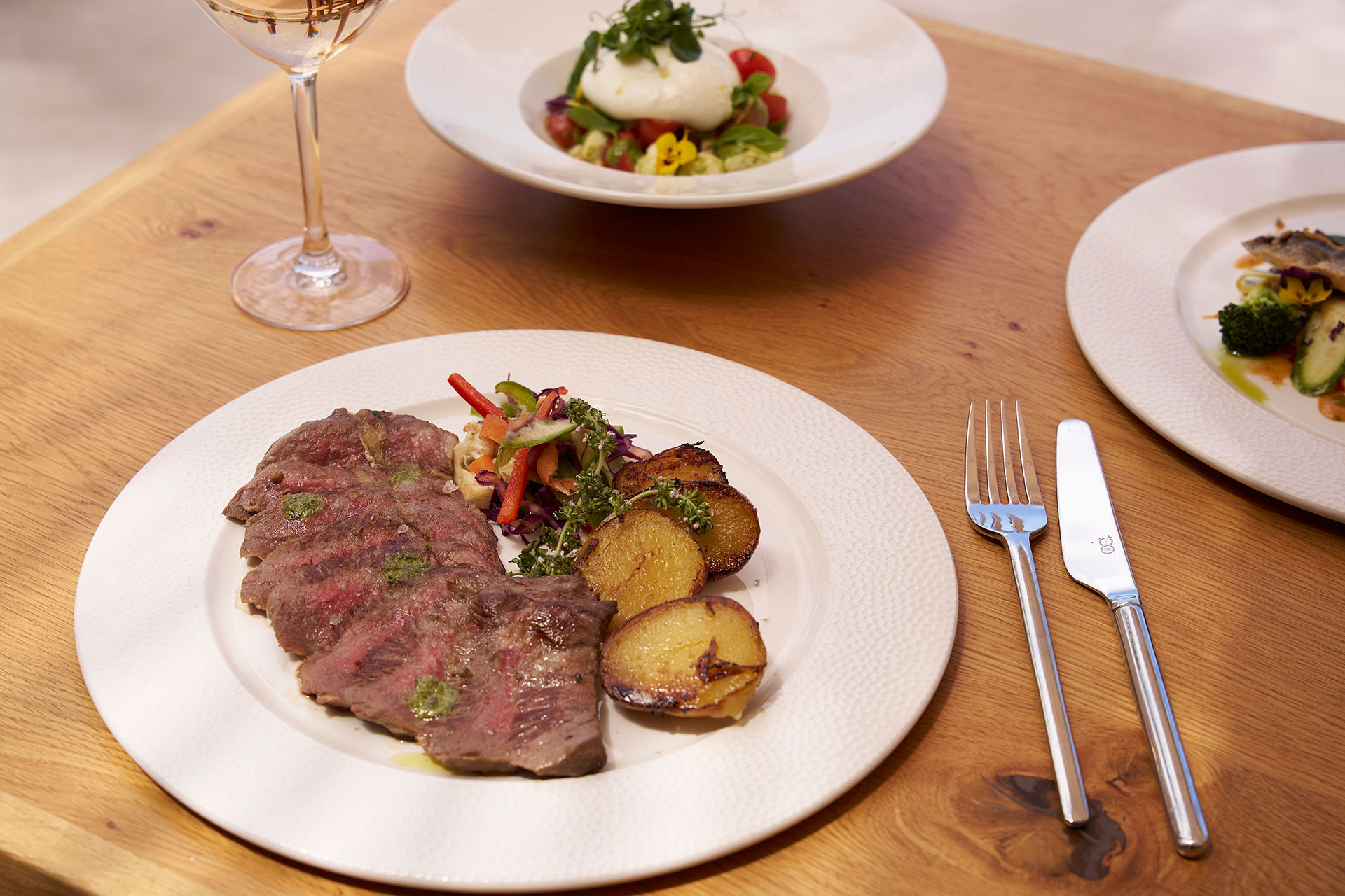 Grilled steak slices served with roasted potatoes and sautéed vegetables on a white plate, accompanied by a glass of wine.