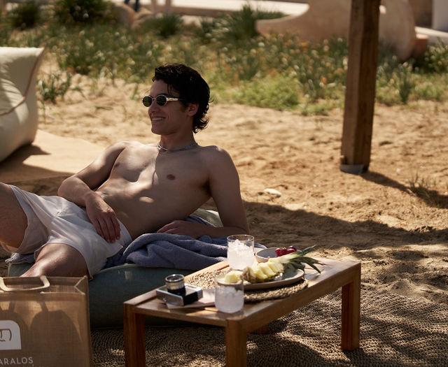 Man relaxing on sunbed at the beach with drinks and fruit on a table.