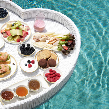 A floating heart-shaped breakfast tray in a pool filled with various dishes and fruit