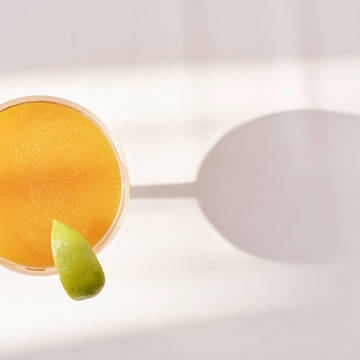 Top-down view of a bright yellow cocktail garnished with a lime wedge, casting a shadow on a white surface.