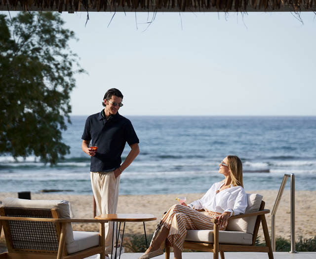 Couple relaxing with drinks by the sea, a man standing and a woman seated on cushioned outdoor chairs facing the ocean.